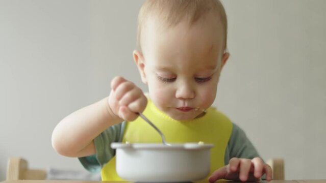 Eating Baby Child Sitting At Table Holding Metal Spoon In Hands Putting Food Into Mouth Chewing On Camera. Portrait Caucasian Toddler Boy Kid Learning To Feed Himself Using Hand And Spoon Yellow Bib