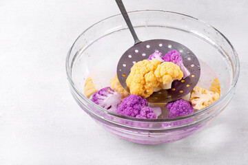 Blanched cauliflower cabbage of purple and yellow colors is cooling down in cold water in glass bowl on white table. Preparing the ingredient