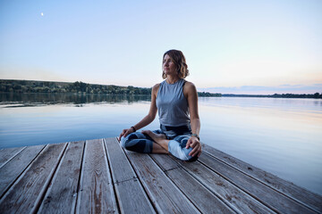 Fototapeta na wymiar A calm yogi woman in a lotus position is meditating on the dock.