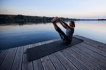 A fit yogi woman is practicing yoga on a dock at dusk. She is in the Boat yoga posture.