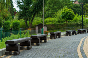 Black wooden benches rhythmically repeating towards the horizon, gray pavement stones, dense greenery of trees in the background, cloudy rainy summer day, deserted place, copy space, selective focus