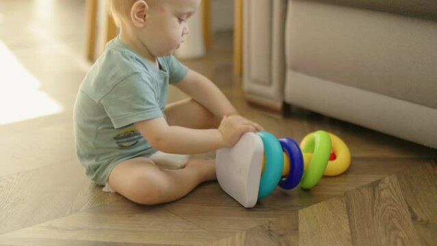 baby child boy kid playing with plastic pyramid sitting on floor in living room. caucasian toddler baby in diaper and green t-shirt learning to collect colorful rings stringing together parts on stick