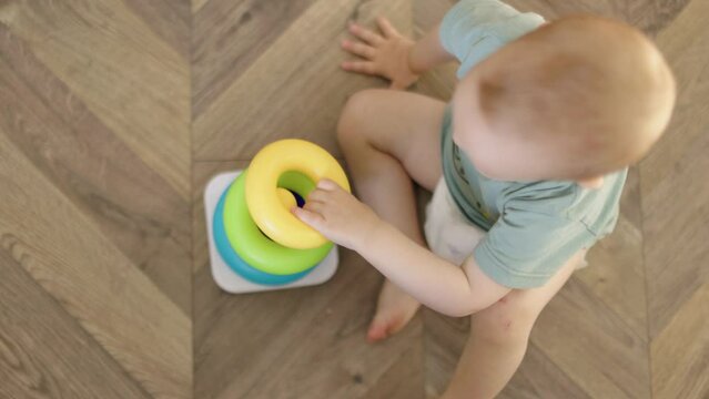 baby child boy kid playing with plastic pyramid sitting on floor in living room. caucasian toddler baby in diaper and green t-shirt learning to collect colorful rings stringing together parts on stick