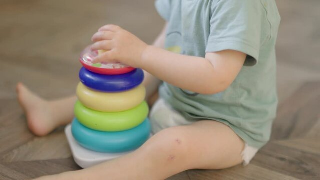 baby child boy kid playing with plastic pyramid sitting on floor in living room. caucasian toddler baby in diaper and green t-shirt learning to collect colorful rings stringing together parts on stick