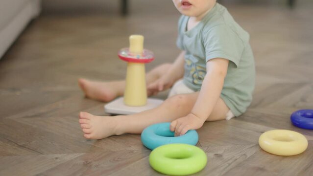 baby child boy kid playing with plastic pyramid sitting on floor in living room. caucasian toddler baby in diaper and green t-shirt learning to collect colorful rings stringing together parts on stick
