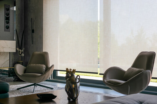 Roller blinds in the interior. Automatic solar shades of large sizes on the window. Fabric with linen texture. In front of a large window is a chair on a carpet. 