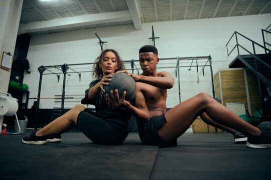 Mixed race friends doing cross fit in the gym. African American male and female sitting down on the floor and passing a medicine ball during exercise routine. High quality photo