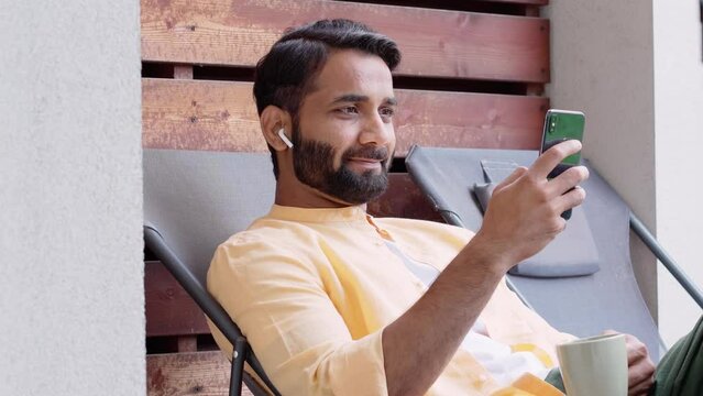 Relaxed indian man sitting outdoor house on chair chilling, drinking coffee, wearing earbud using smartphone looking at cell phone watching video news, checking social media, listening music podcast.