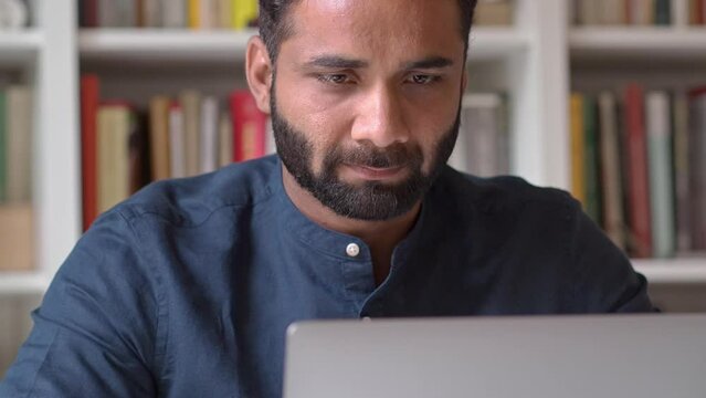 Busy serious indian business man manager remote working or learning online looking at laptop sitting in office at home using software on computer thinking on question analyzing market data technology.