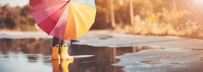 Child standing in the puddle in yellow rubber boots and holding colourful umbrella in hands
