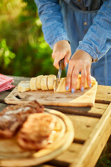 Fototapeta Unrecognizable woman cutting bread for bruschetta on wooden board during weekend barbecue in yard, outdoor, prepare for grilling, summer family picnic, food on the nature. obraz