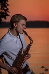 A young guy plays the saxophone in the evening at sunset.