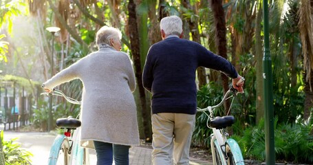 Healthy, active and retired senior couple enjoying a walk with their bicycles through a park with lens flare from behind. Happy and loving elderly man and woman talking and enjoying a stroll outdoors - Powered by Adobe