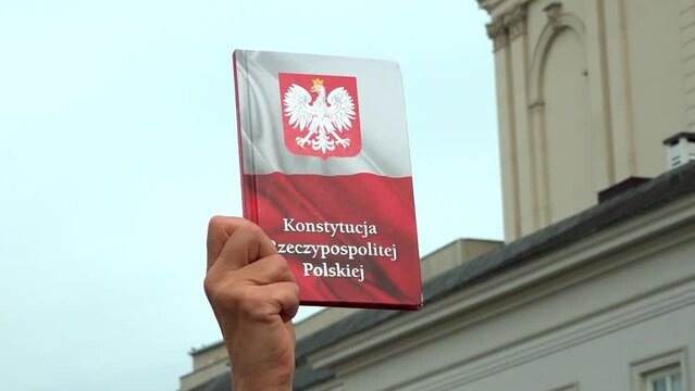 A hand holds a book of the Constitution of Poland close-up against the sky. Translation: The Constitution of the Republic of Poland.