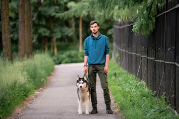 Naklejka na ściany i meble A cute young man is walking along the road with his beloved Siberian Husky dog in nature.