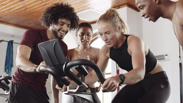 Diverse group of fit young adults cheering around Caucasian female friend while she cycles on stationary exercise bike at an indoor fitness gym.