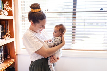Young smiling mum with hair in a bun holding young baby inside home