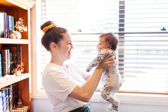 Lifestyle image of young mother playing with newborn baby inside home with sunlight shining in
