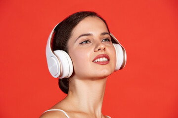 Fototapeta premium Close-up portrait of beautiful young woman posing in white headphones isolated over red studio background