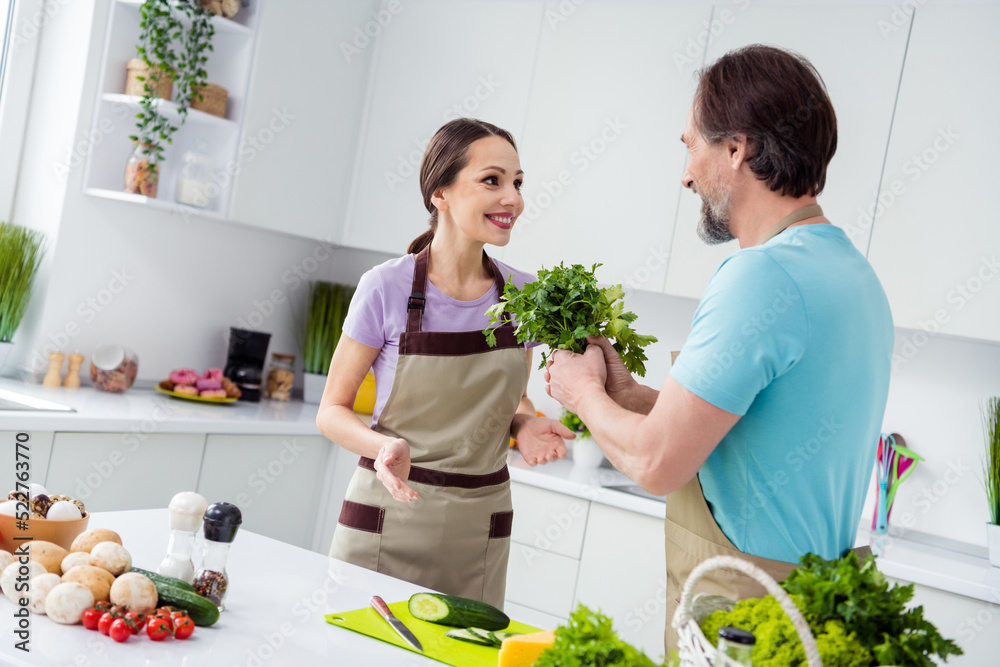 Wall mural Portrait of two cheerful satisfied partners hold fresh organic vegetable parsley enjoy cooking free time indoors