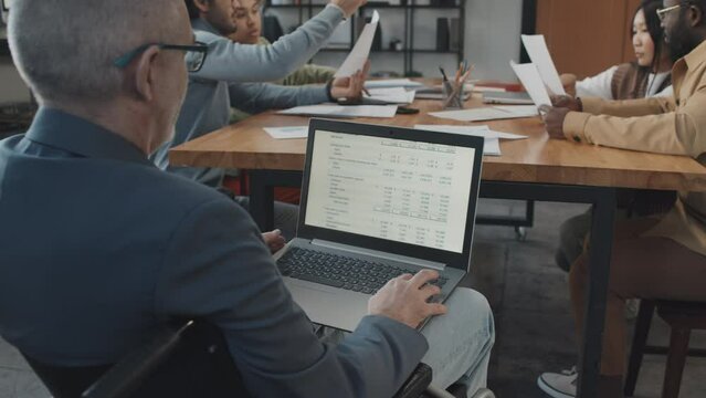 Over-shoulder of senior man in wheelchair using laptop computer on business meeting in conference room in morning