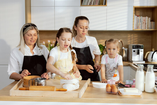 Grandmother Mom And Daughters Cooking Baking Cake Older Daughter Taking Flour Pouring Into Vessel Mother Giving Orders Preschooler Daughter. Granny Watching Process. Family Having Fun.