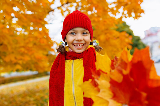 A girl with a wide smile is playing with red and yellow leaves in an autumn park.