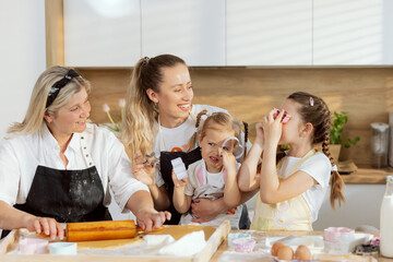 Happy granddaughters looking at mother and granny holding cookies cutters as glasses laughing. Elderly grandmother rolling dough looking at girls. Happy family having fun baking cooking in kitchen.