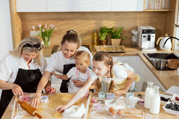Curious daughters helping eldely granny and happy young mother preparing christmas cookies for baking. Family having fun together in modern light pretty kitchen. Preparing surprise for dinner.