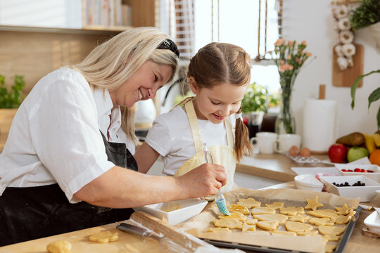 Beautiful mother with grey hair in the kitchen teaching curious daughter oiling cookies preparing before putting in oven. Having fun together at weekend baking cookies biscuits for breakfast.