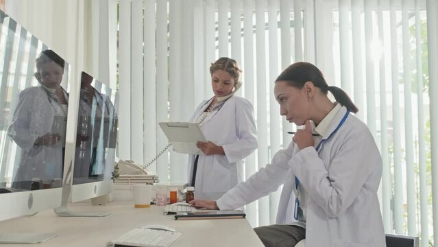 Female doctor working with x ray image on computer as her colleague having phone talk and taking notes on clipboard in clinic