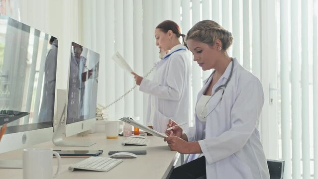 Female doctor sitting at desk, examining x ray image on computer and taking notes on clipboard as her colleague speaking on phone during workday in medical office