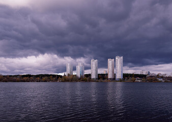Fototapeta premium View across the dark lake to tall new houses in the city near nature. Fantasy nature. Dark cloudy landscape. Dark terrible clouds