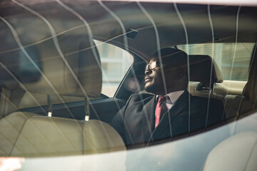 A dark-skinned businessman in a suit and glasses in the back seat of a luxury car looks out the window. Premium confidence and comfort