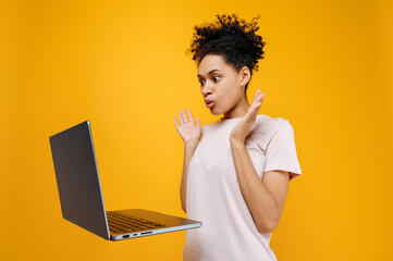 Emotional puzzled shocked curly haired african american young woman, looks in surprise at the laptop screen, gesticulates with her hands, saw the unexpected news, stands on isolated orange background