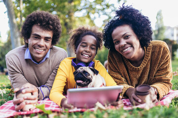 Beautiful young family lying on a picnic blanket with their dog, enjoying an autumn day in park while using digital tablet.