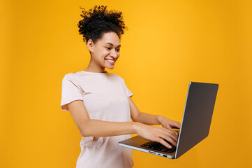 Happy excited african american young woman with curly hair, looks at laptop screen, saw unexpected good information, great news, stands over isolated orange background, emotional facial expression