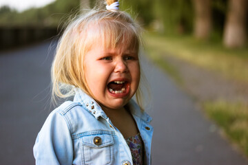 Little blonde hysteric girl stands in a middle of a street crying out loud with open mouth. Poor child is lost. Real negative emotions, anguish, crankiness, horror concept. Abandoned child, female kid