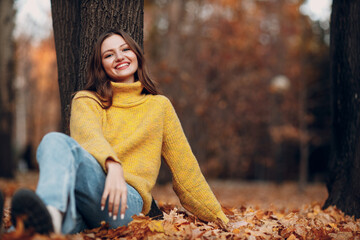 Beautiful smiling girl at autumn park sitting fall yellow foliage leaves.