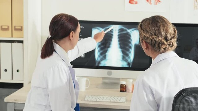 Back view of two female doctors discussing chest x ray scan on computer screen while cooperating during workday in clinic