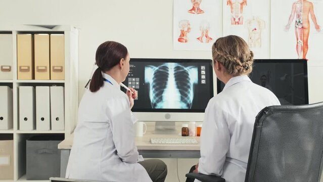 Back view of two female doctors examining chest x ray scan on computer and discussing diagnosis while working together in clinic