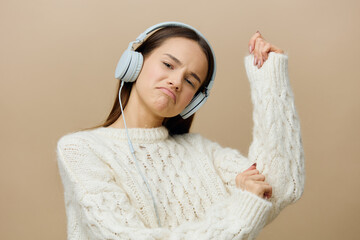 a happy, joyful woman stands on a light gray background in a knitted sweater and joyfully raises her hands up with happiness, listening to music with headphones. 