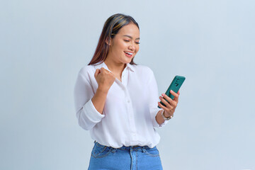 Excited young Asian woman looking at mobile phone screen gesturing yes with raised fist reacting to online news isolated over white background - obrazy, fototapety, plakaty