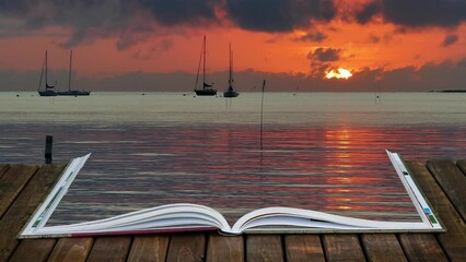 Open book with beautiful sunrise near water edge with boats in distance