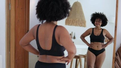 A beautiful African-American woman in black lingerie looks at her sexy curvaceous forms in front of a mirror. The concept of body positivity. Multiracial woman in front of a mirror.
