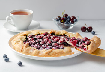 Fresh cherry and blueberry galette with coconut on plate with a cup of tea, white background. Homemade rustic dessert - american pie or tart or cake or french galette
