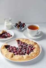 Fresh cherry and blueberry galette with coconut on plate with a cup of tea, white background, copy space. Homemade rustic dessert - american pie or tart or cake or french galette