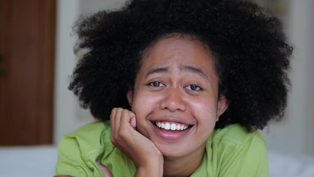 A happy black woman close-up with an afro hairstyle lies on the bed and smiles with a snow-white smile and sings songs. Portrait of afro asian woman in bedroom on bed home interior.