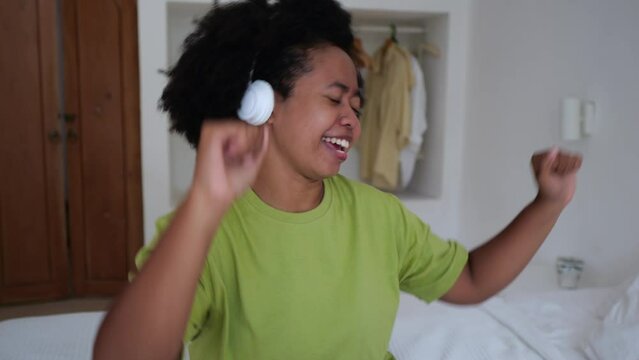 A black woman sings a song on the bed enjoys listening to music on headphones in the bedroom. A mixed race woman with an Afra hairstyle dances to music from her headphones.
