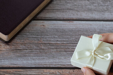 Female hand holding a wrapped gift box with a ribbon on a wooden background with a closed Holy Bible Book. Copy space. A closeup.